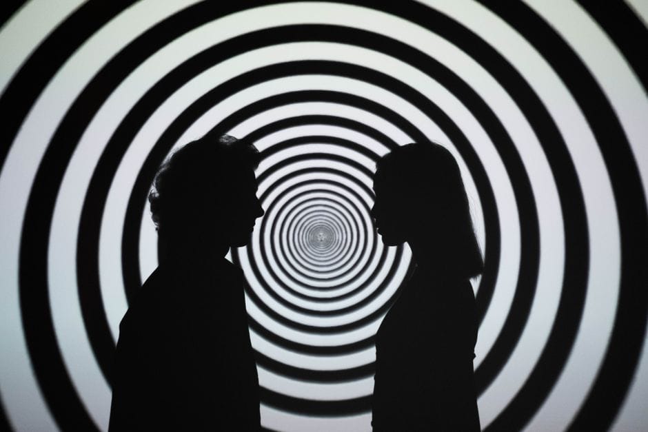 Silhouette zweier Personen, die sich gegenüberstehen, vor einem hypnotischen Spiralhintergrund, der eine optische Täuschung erzeugt. KI Halluzination